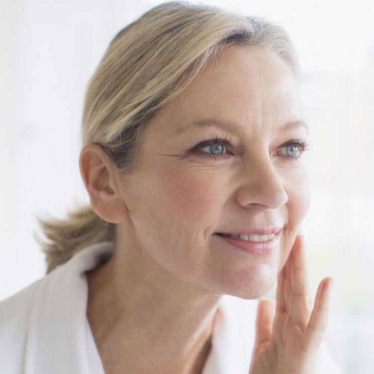 Mulher madura sorrindo suavemente ao tocar o rosto, em momento de autocuidado e bem-estar, com luz natural ao fundo.