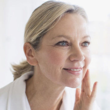Mulher madura sorrindo suavemente ao tocar o rosto, em momento de autocuidado e bem-estar, com luz natural ao fundo.