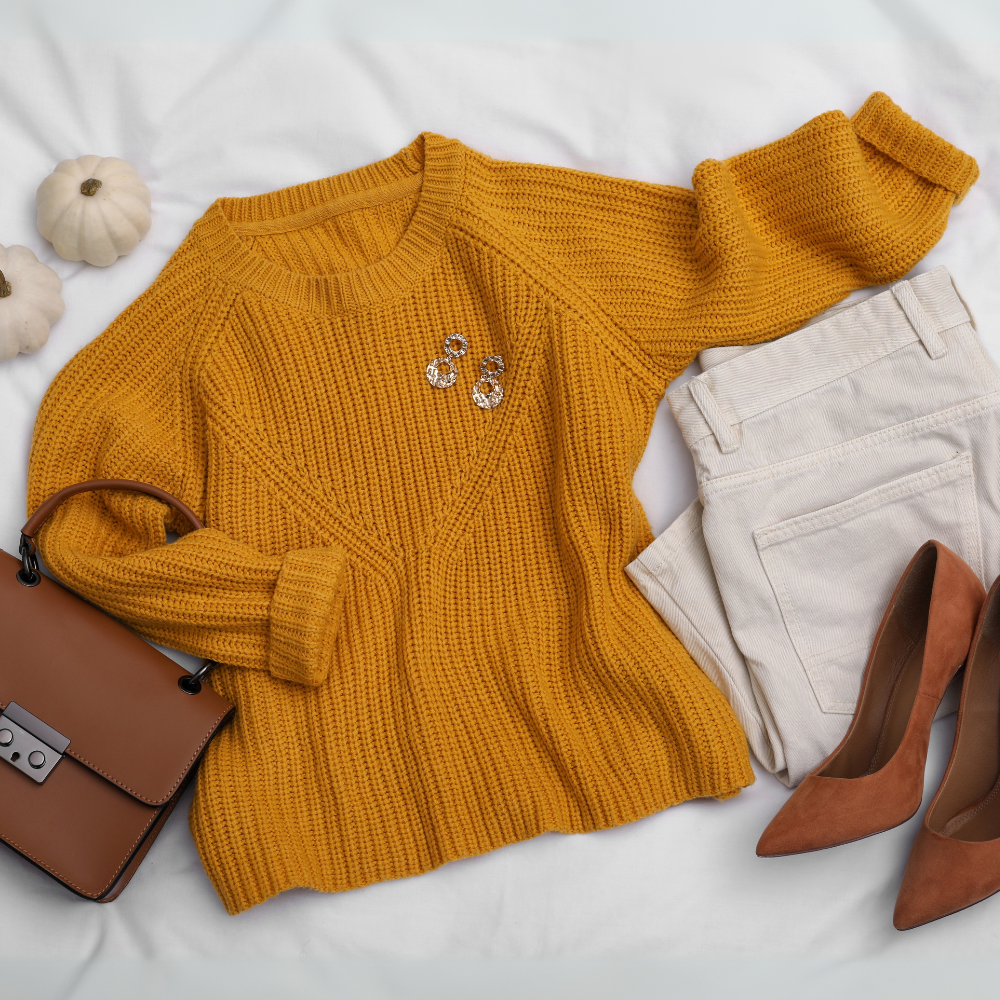 Look outonal com suéter mostarda de tricô, calça off-white de sarja, sapato caramelo de salto, bolsa marrom e brincos dourados sobre fundo branco.