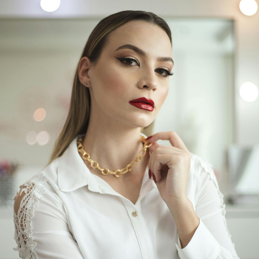Mulher com maquiagem marcante e batom vermelho, usando camisa branca com detalhes rendados e colar dourado, transmitindo elegância e confiança.
