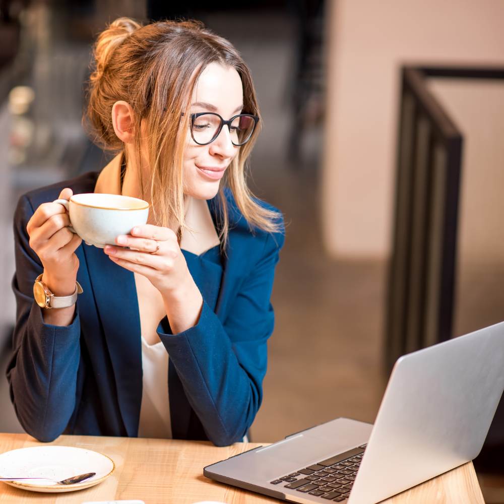 Mulher sorridente de blazer azul tomando café em frente ao notebook, transmitindo leveza e bem-estar durante o trabalho.