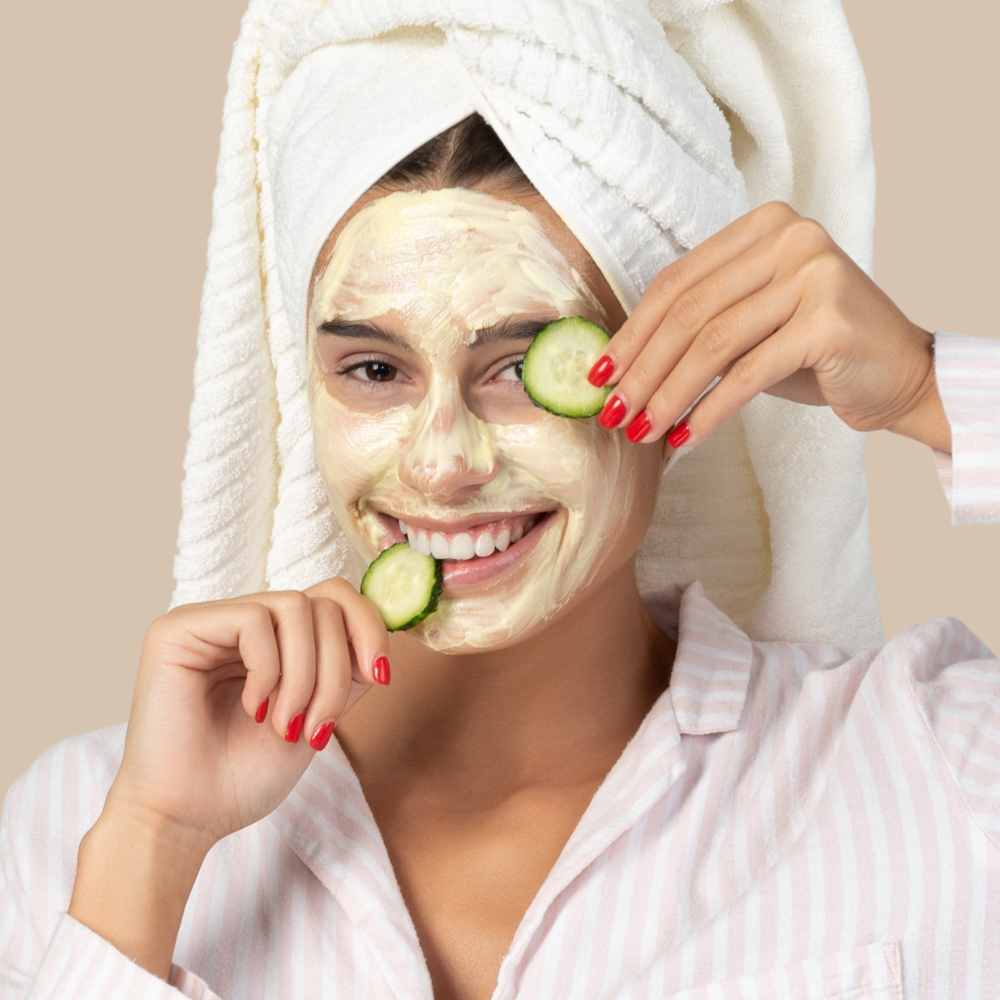 Mulher com toalha na cabeça, máscara facial e pepinos nos olhos, sorrindo em momento divertido de skincare e autocuidado.