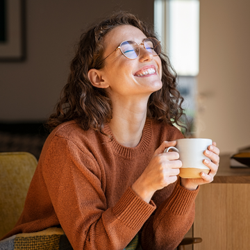 Mulher sorrindo com os olhos fechados enquanto segura uma caneca, vestindo suéter marrom, em momento de bem-estar e pausa aconchegante.