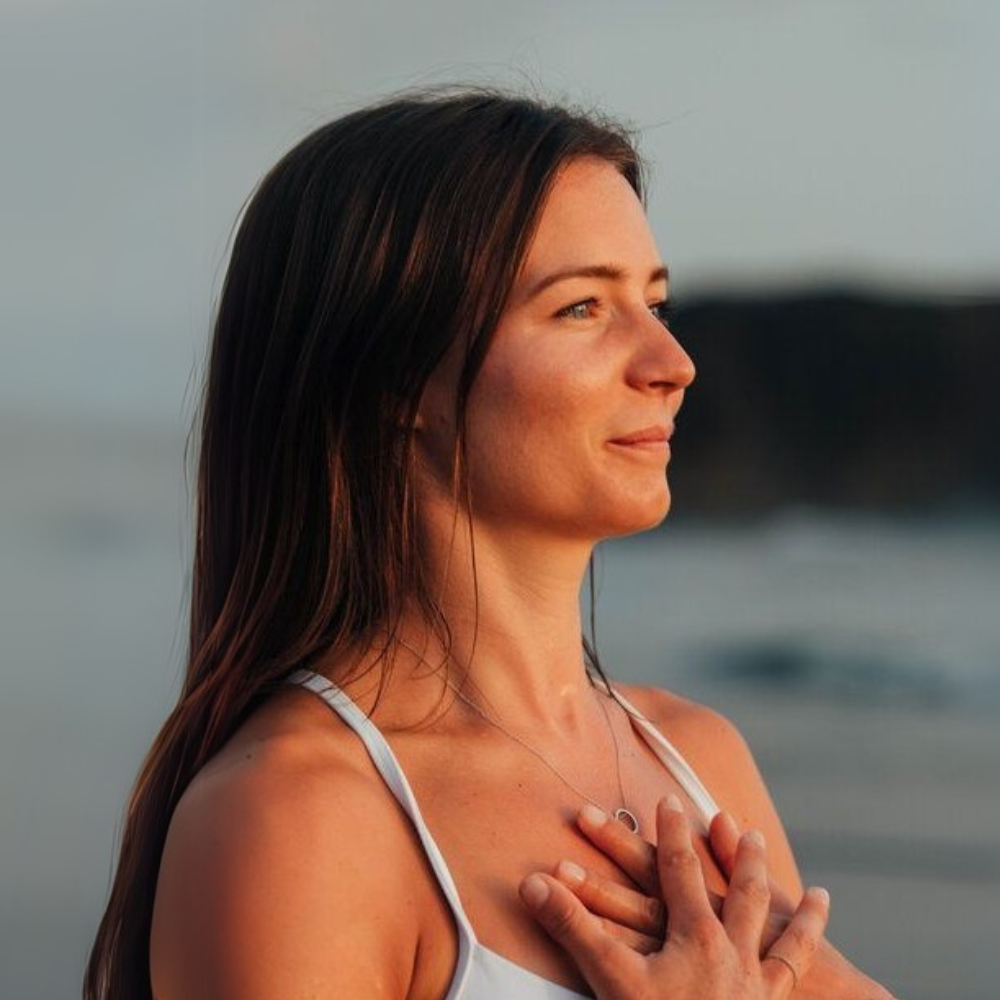 Mulher de olhos fechados ao ar livre com as mãos no peito, em momento de respiração e autocuidado, à beira-mar durante o pôr do sol.