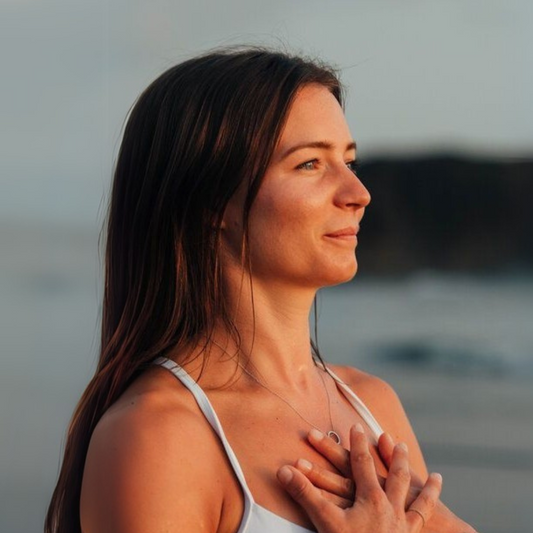Mulher de olhos fechados ao ar livre com as mãos no peito, em momento de respiração e autocuidado, à beira-mar durante o pôr do sol.