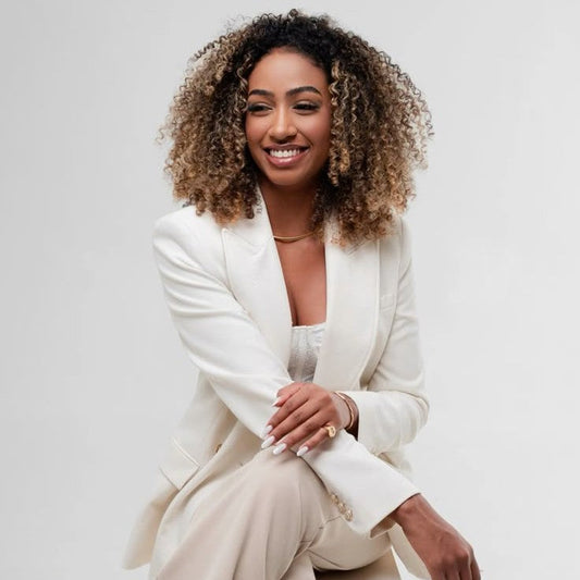 Mulher sorridente com blazer branco e look elegante, sentada em pose confiante, com fundo claro e estilo sofisticado.