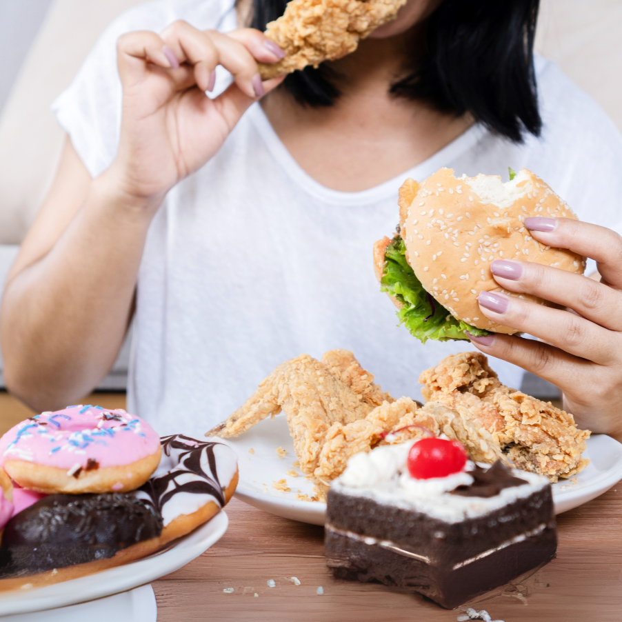 Pessoa segurando hambúrguer e frango frito à mesa, cercada por doces como bolo e donuts, representando episódio de alimentação emocional.