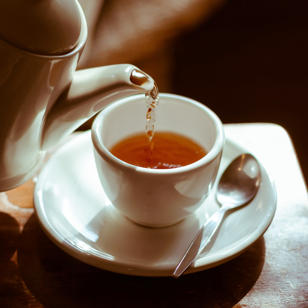 Xícara branca com chá quente sendo servido por um bule, em uma mesa de madeira com colher ao lado — cena acolhedora e relaxante que remete ao bem-estar.