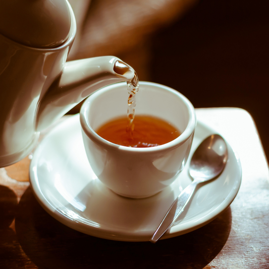 Xícara branca com chá quente sendo servido por um bule, em uma mesa de madeira com colher ao lado — cena acolhedora e relaxante que remete ao bem-estar.
