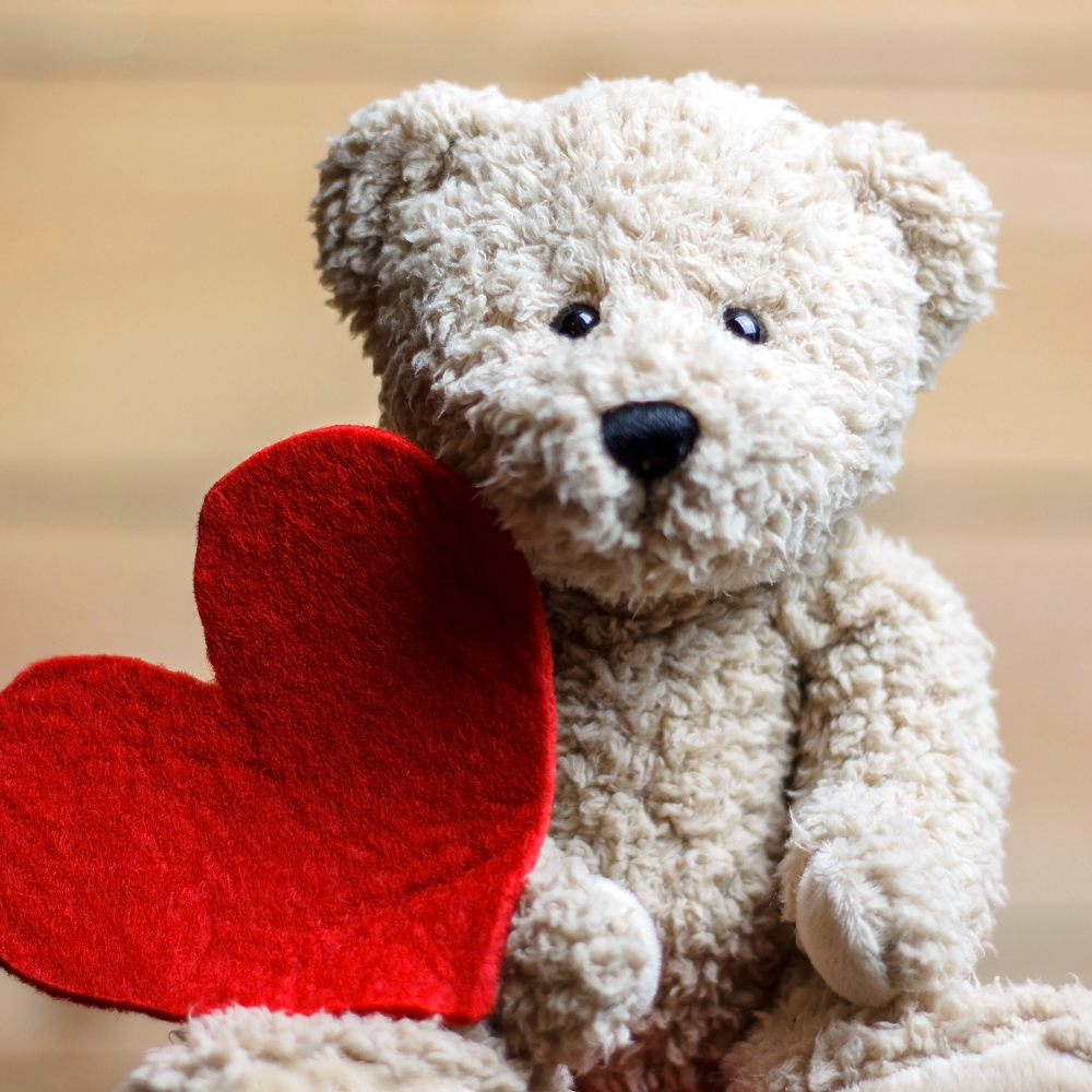 Urso de pelúcia bege segurando um coração vermelho de feltro, em fundo desfocado de madeira clara, simbolizando carinho e afeto.
