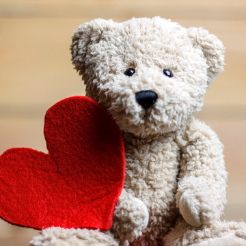 Urso de pelúcia bege segurando um coração vermelho de feltro, em fundo desfocado de madeira clara, simbolizando carinho e afeto.