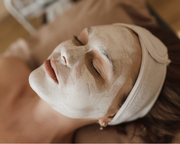 Mulher deitada com máscara facial branca e faixa na cabeça, aproveitando um momento de cuidado e hidratação para a pele.