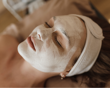 Mulher deitada com máscara facial branca e faixa na cabeça, aproveitando um momento de cuidado e hidratação para a pele.