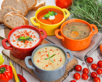Variedade de sopas coloridas e quentinhas servidas em panelinhas, acompanhadas de pães, tomates e ervas frescas, em uma mesa rústica.