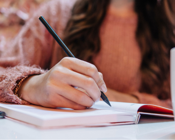 Mulher escrevendo em um caderno em ambiente tranquilo, simbolizando revisão e planejamento de metas pessoais.