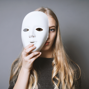 Mulher segurando uma máscara branca à frente do rosto, simbolizando o sentimento de esconder sua verdadeira identidade. Representação visual da síndrome da impostora.