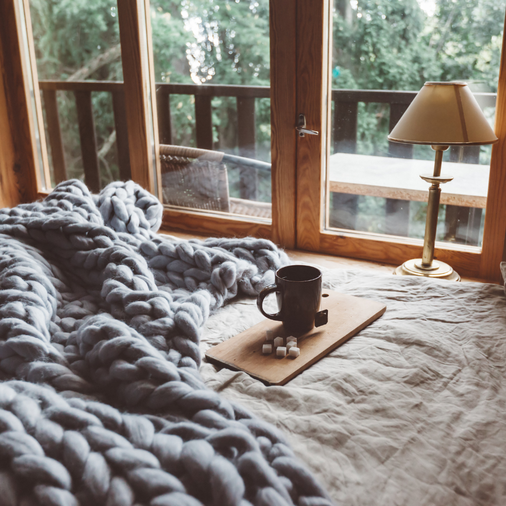Cama com manta grossa e xícara de café ao lado de janela com vista para a natureza, transmitindo aconchego e bem-estar em casa.