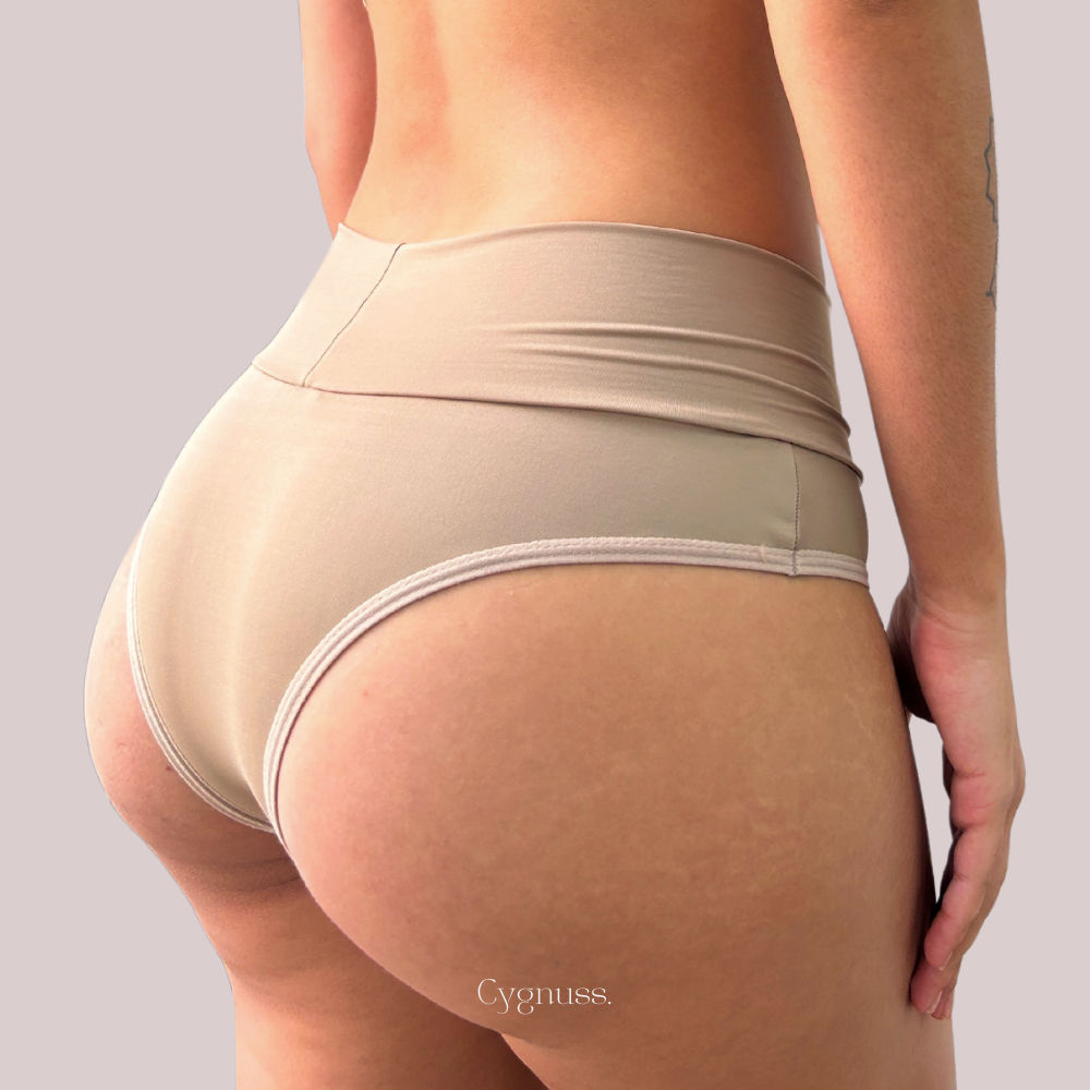 Mulher usando Calcinha Pandora, tanga de cintura média, toque suave, conforto, cor nude, marca Cygnuss, vista traseira.