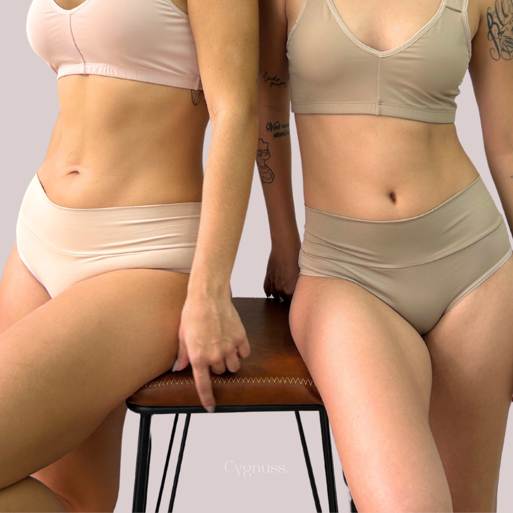 Duas mulheres usando calcinhas Cygnuss Pandora de cintura média, exibindo conforto e estilo, sentadas em um banco em tons neutros.