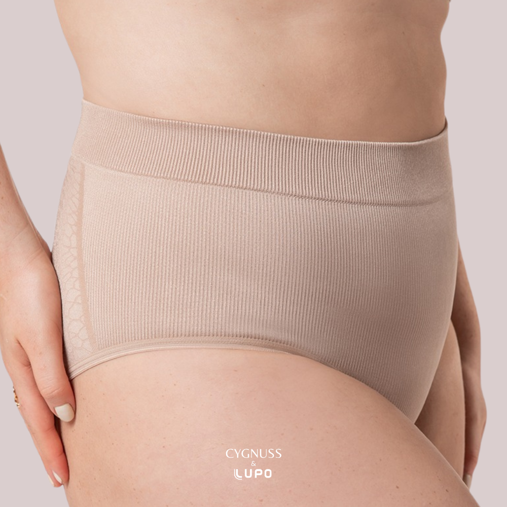 Close-up de uma mulher usando uma calcinha modeladora nude de cintura alta. O tecido texturizado e sem costuras proporciona compressão suave e conforto, modelando a silhueta. Detalhes laterais com acabamento diferenciado. O logotipo da Cygnuss & Lupo está sobreposto na parte inferior da peça. Fundo nude rosado.