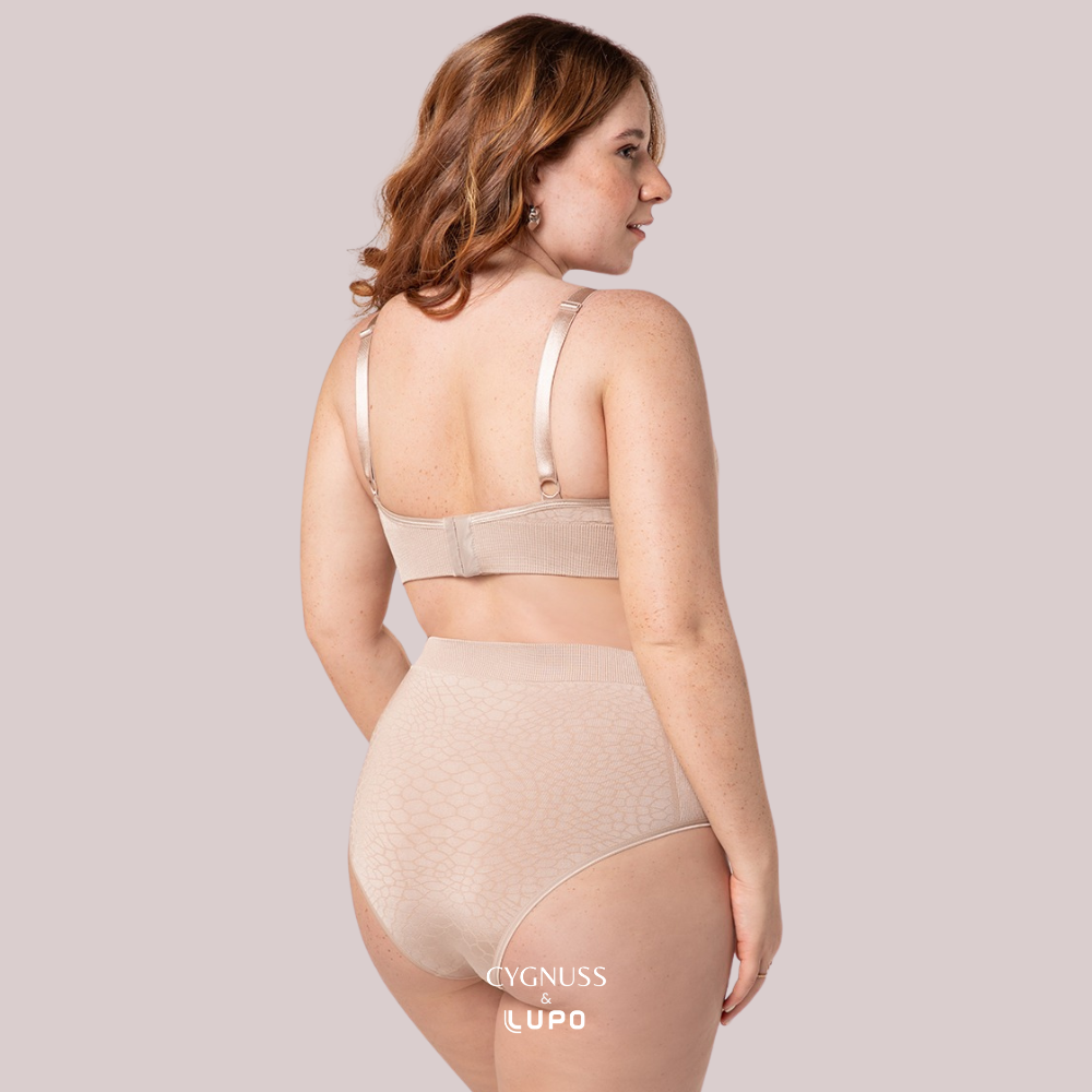Vista traseira de uma mulher ruiva de cabelos cacheados usando um conjunto de lingerie bege sem costuras, composto por um sutiã modelador com fecho traseiro e uma calcinha de cintura alta. O tecido texturizado proporciona ajuste perfeito, sustentação e conforto. A modelo sorri suavemente, transmitindo segurança e bem-estar. O logotipo da Cygnuss & Lupo está sobreposto na parte inferior da peça.