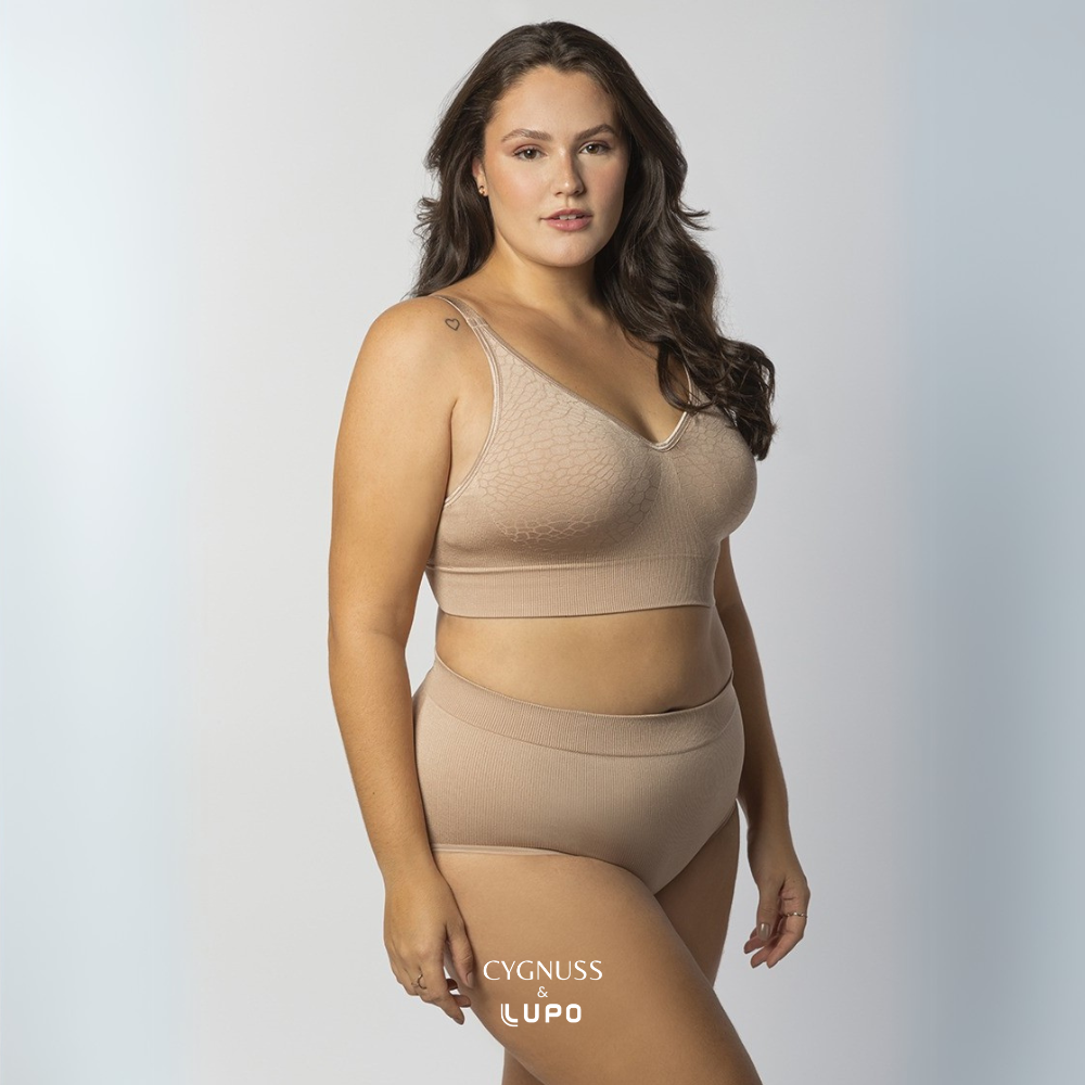 Mulher com corpo curvilíneo e cabelos longos usando um conjunto de lingerie nude sem costuras, composto por um sutiã modelador e uma calcinha de cintura alta. O tecido texturizado e elástico proporciona ajuste perfeito, conforto e sustentação. O logotipo da Cygnuss & Lupo está sobreposto na parte inferior da peça. Fundo branco com leve degradê azul.