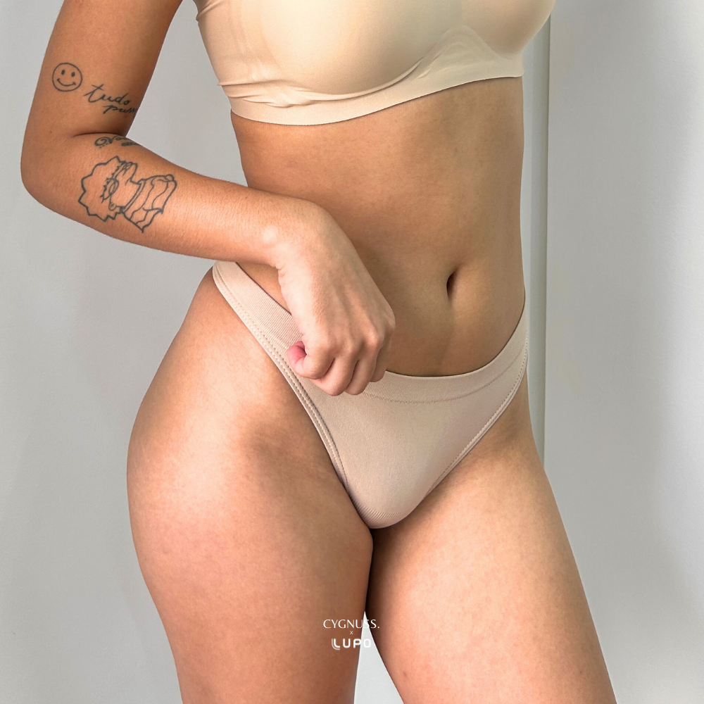 Close frontal da calcinha fio Cygnuss x Lupo na cor bege, vestida por modelo tatuada. A peça em microfibra tem laterais médias, toque macio e acabamento sem costura, garantindo conforto, leveza e um visual discreto sob qualquer roupa.