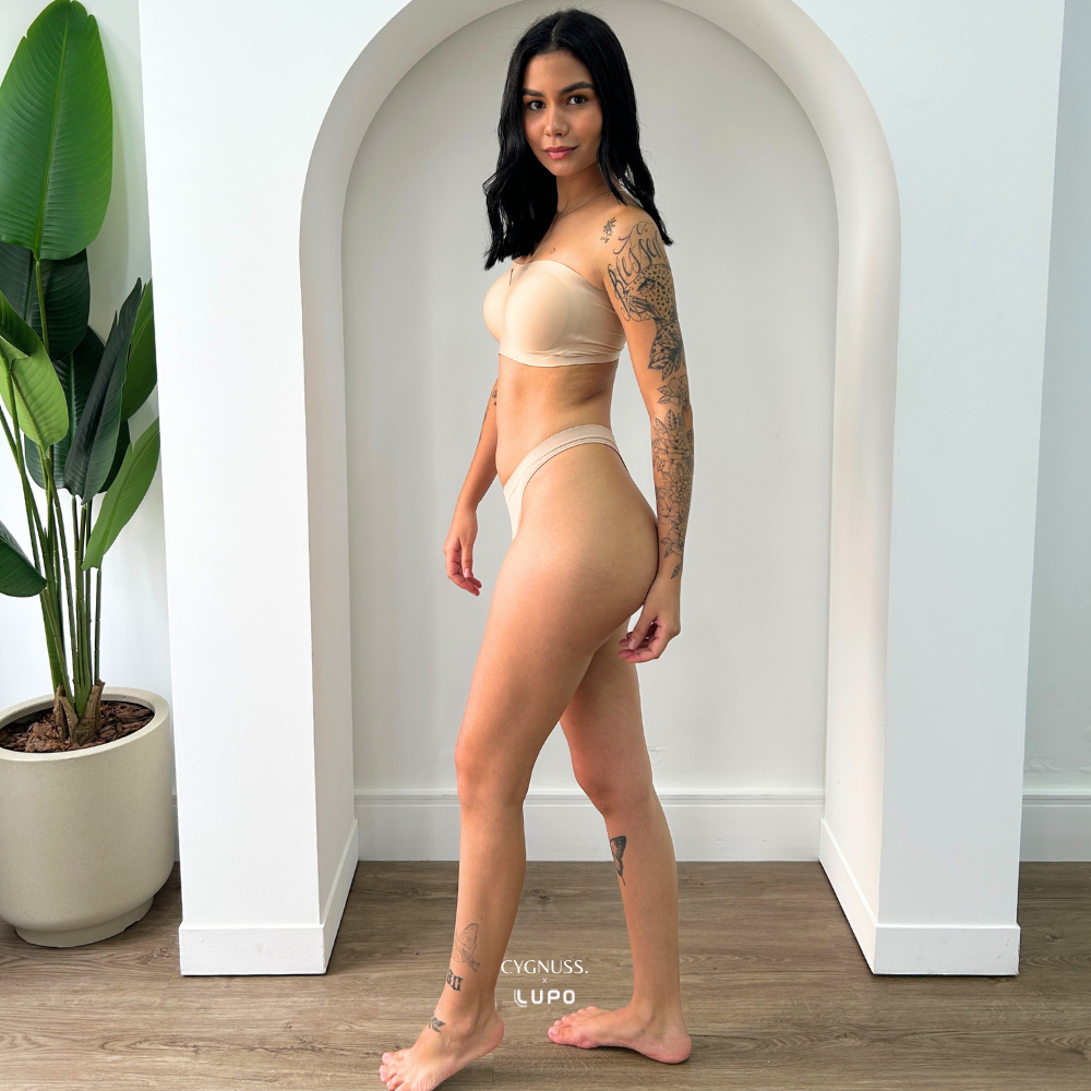 Modelo tatuada em vista lateral vestindo calcinha fio bege Cygnuss x Lupo. A peça em microfibra possui toque macio, laterais médias e acabamento sem costura, oferecendo ajuste perfeito e discreto sob a roupa. Ideal para quem busca leveza com sensualidade.