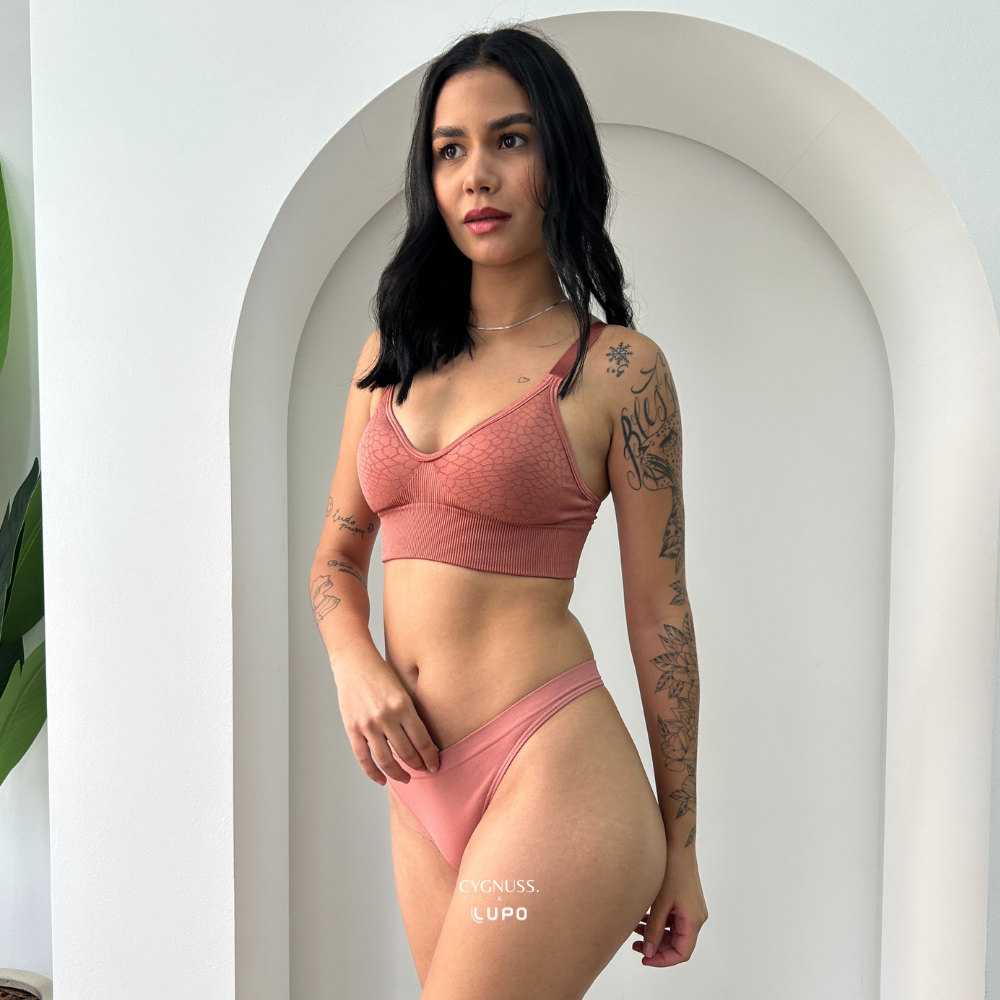 Close frontal da calcinha fio Cygnuss x Lupo na cor rosé, vestida por modelo tatuada. A peça é feita em microfibra macia, com laterais médias e acabamento sem costuras que proporciona conforto invisível sob a roupa. Ideal para quem busca leveza, liberdade e um caimento que valoriza as curvas sem marcar.