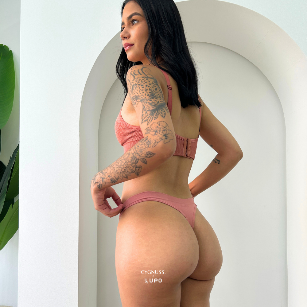 Destaque para a Calcinha Microfibra Fio Cygnuss x Lupo na cor rosé. A modelo aparece de costas, evidenciando o corte fio dental que valoriza o bumbum com conforto. O tecido em microfibra se ajusta perfeitamente ao corpo, com acabamento sem costuras e toque macio.