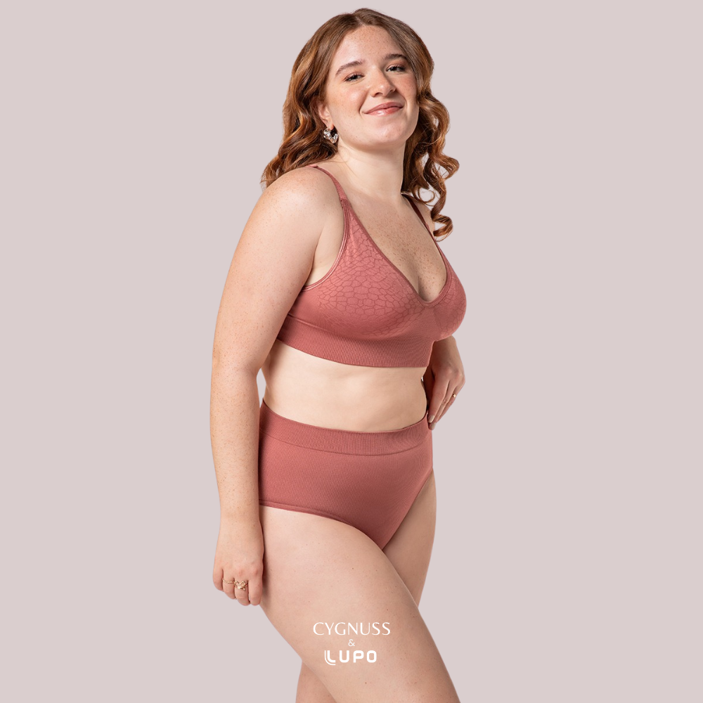 Mulher ruiva de cabelos cacheados usando um conjunto de lingerie na cor telha, composto por um sutiã sem costuras e uma calcinha modeladora de cintura alta. O tecido texturizado oferece ajuste natural, sustentação e conforto ao longo do dia. A modelo sorri de forma leve, transmitindo bem-estar e segurança. O logotipo da Cygnuss & Lupo está sobreposto na parte inferior da peça.