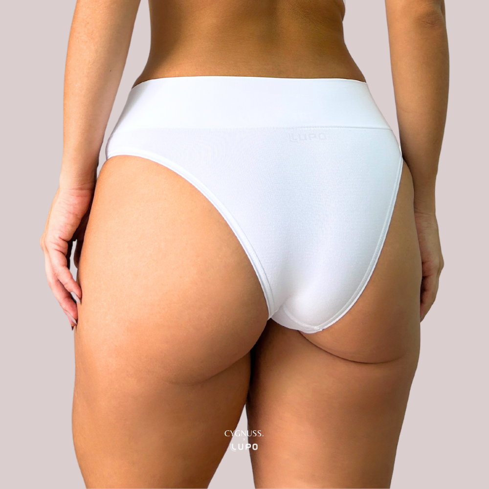Vista traseira de uma mulher usando calcinha modeladora branca da marca Lupo em parceria com a Cygnuss. A peça tem cós alto, acabamento sem costura e modelagem anatômica. Fundo lilás claro.