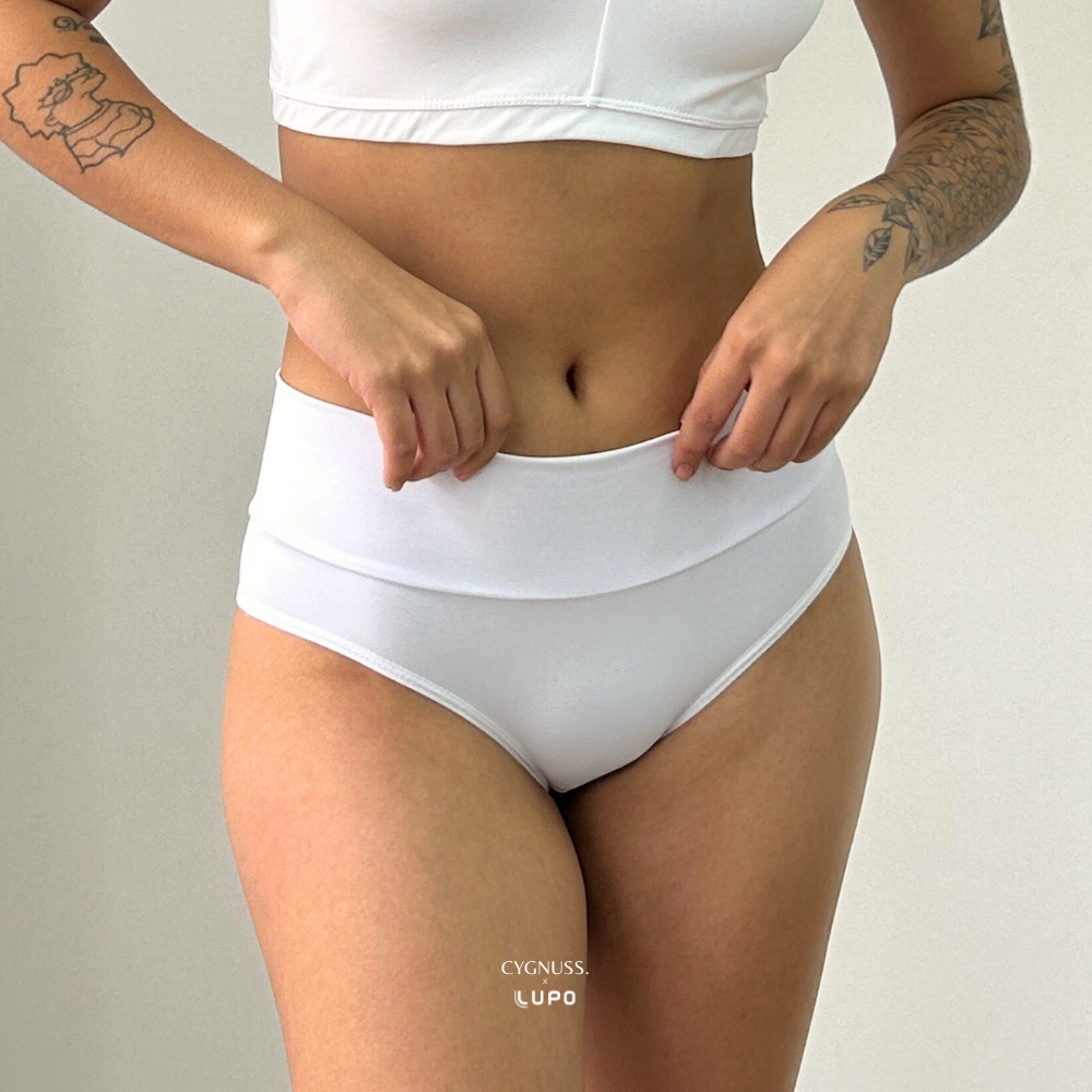 Mulher vestindo top e calcinha modeladora brancos da parceria Cygnuss + Lupo, ajustando a peça com as mãos. A calcinha tem cintura alta e tecido ajustado ao corpo. Fundo branco minimalista.
