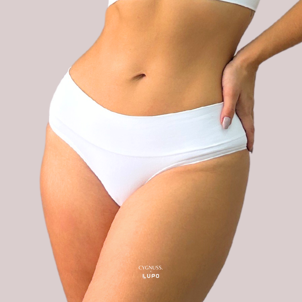Mulher usando calcinha modeladora branca da marca Lupo em parceria com a Cygnuss. A peça tem cós alto, modelagem anatômica e tecido sem costura. Fundo lilás claro.