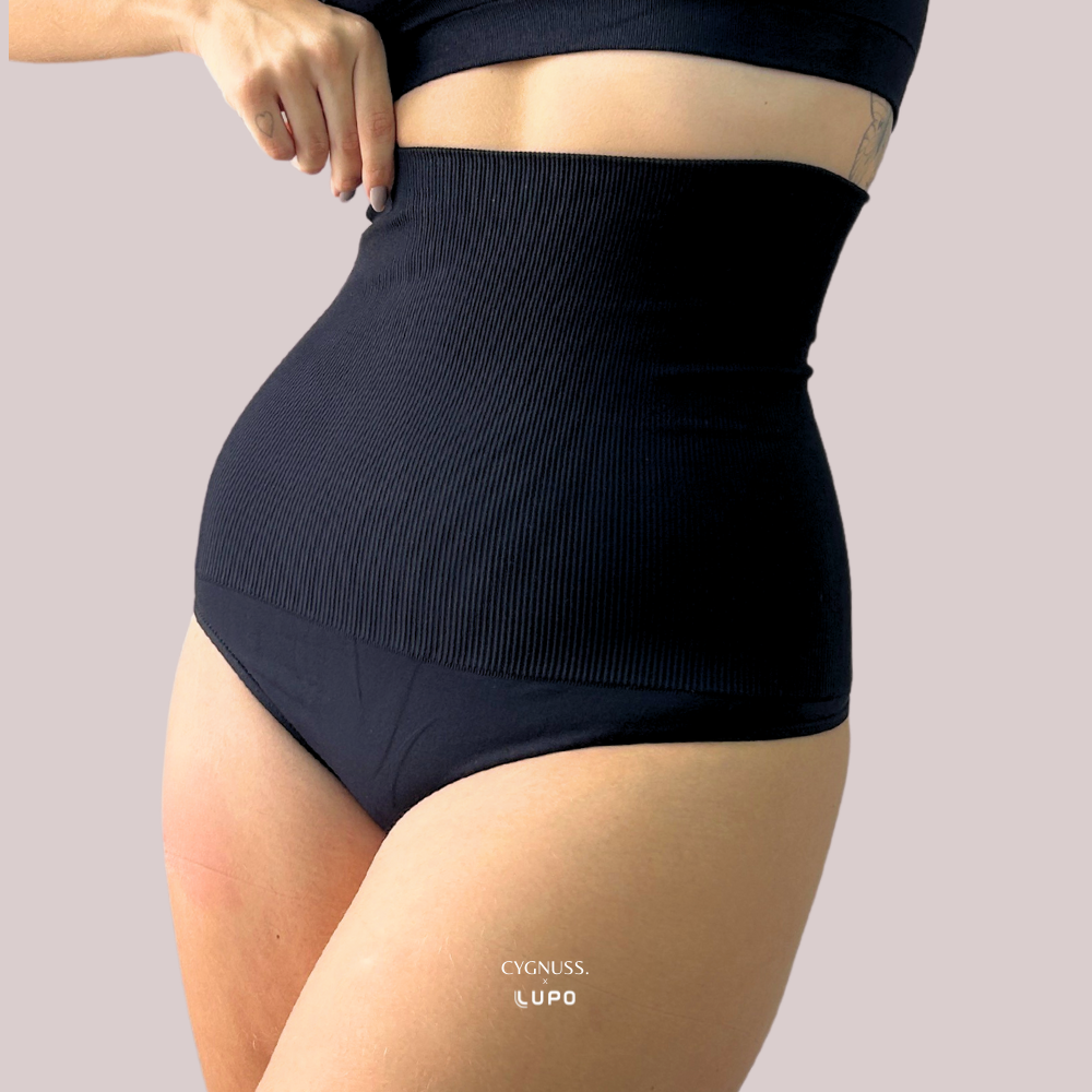 Close-up de uma mulher vestindo uma calcinha modeladora preta de cintura alta, ajustando a peça na região abdominal. O tecido texturizado proporciona compressão e modelagem ao corpo, garantindo um ajuste firme e confortável. O logotipo da Cygnuss & Lupo está sobreposto na parte inferior da peça.