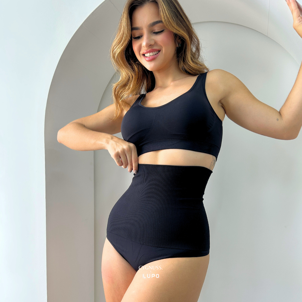 Mulher sorridente vestindo um conjunto preto de top e calcinha modeladora de cintura alta com compressão. A peça tem tecido canelado sem costuras, proporcionando conforto e ajuste perfeito ao corpo. A modelo puxa levemente a cintura da calcinha, destacando sua elasticidade e efeito modelador. O logotipo da Cygnuss & Lupo está sobreposto na parte inferior da peça. Fundo minimalista com arco branco.