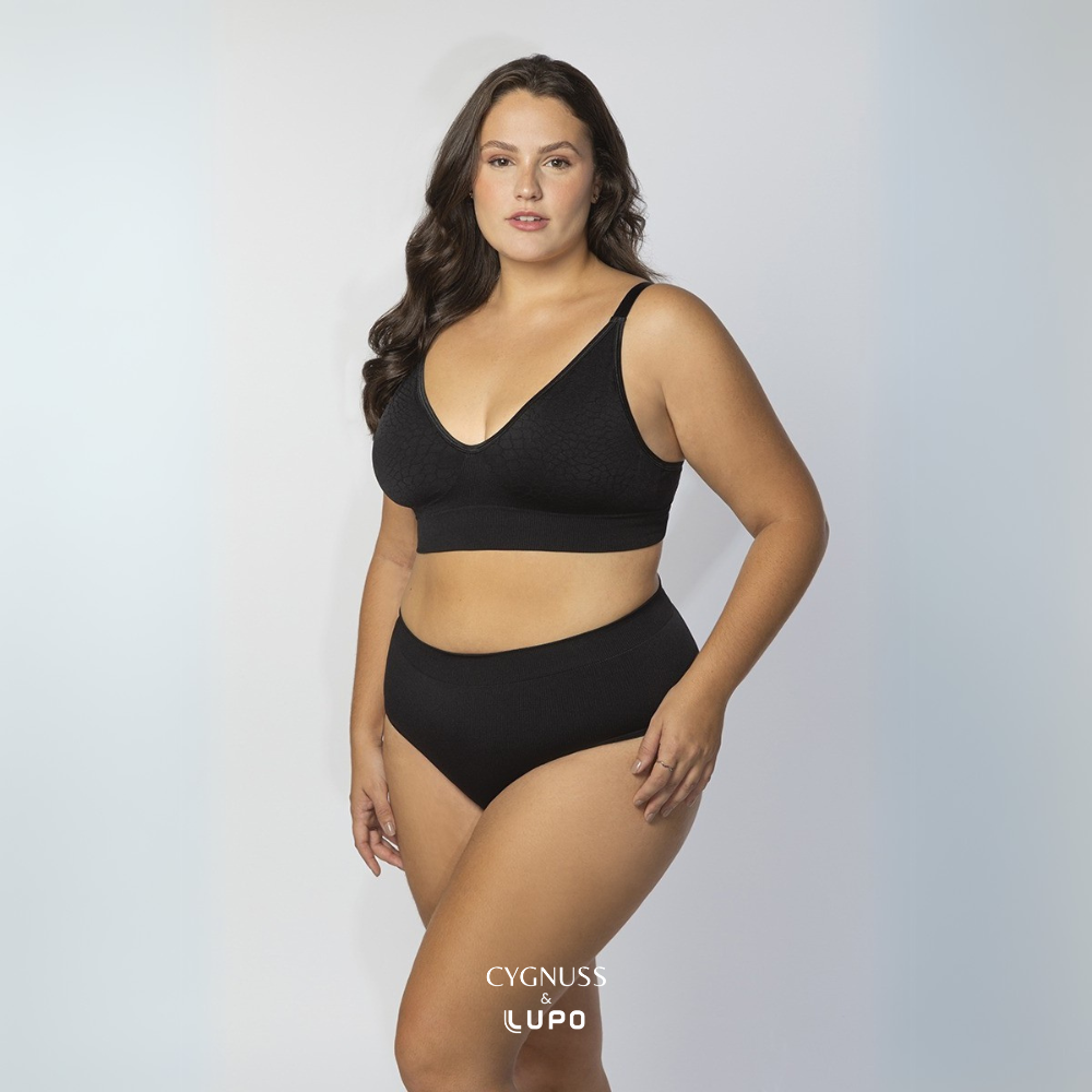 Mulher com corpo curvilíneo e cabelos longos usando um conjunto de lingerie preto sem costuras, composto por um sutiã modelador e uma calcinha de cintura alta. O tecido texturizado proporciona ajuste perfeito, sustentação e conforto total ao longo do dia. O logotipo da Cygnuss & Lupo está sobreposto na parte inferior da peça.