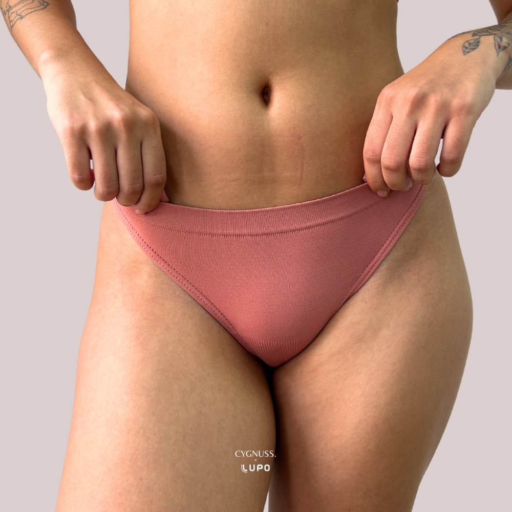 Close frontal de mulher vestindo calcinha modeladora rosé, cintura alta e sem costuras aparentes. A peça realça as curvas com conforto e leve compressão, com logo Cygnuss e Lupo na parte inferior da imagem.