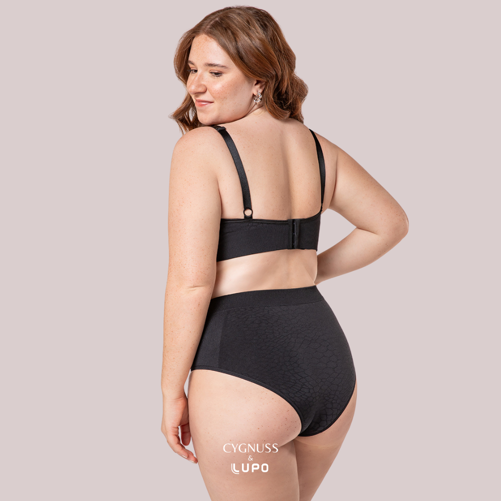 Vista traseira de uma mulher ruiva de cabelos cacheados usando um conjunto de lingerie preto sem costuras, composto por um sutiã modelador com fecho traseiro e uma calcinha de cintura alta. O tecido texturizado proporciona ajuste perfeito, sustentação e conforto. A modelo sorri suavemente, transmitindo segurança e bem-estar. O logotipo da Cygnuss & Lupo está sobreposto na parte inferior da peça.