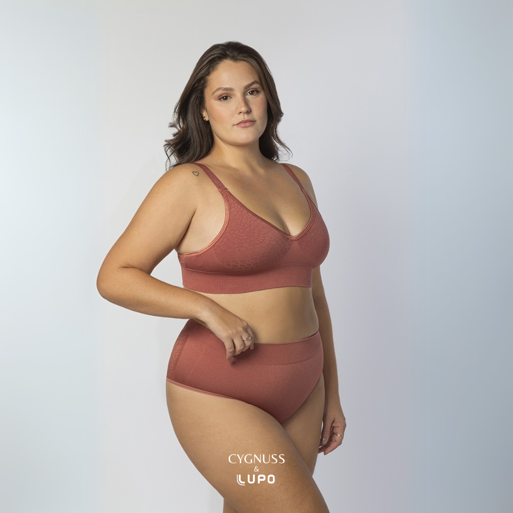 Mulher com corpo curvilíneo e cabelos longos usando um conjunto de lingerie na cor telha, composto por um sutiã modelador e uma calcinha de cintura alta. O tecido texturizado sem costuras oferece sustentação, conforto e modelagem ao corpo. A modelo posa com uma mão na cintura, destacando o ajuste perfeito das peças. O logotipo da Cygnuss & Lupo está sobreposto na parte inferior da peça.