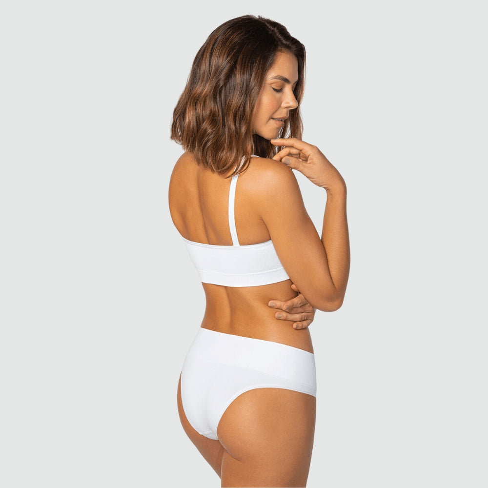 Modelo de costas usando conjunto branco da Cygnuss + Lupo, com top de alças finas e calcinha cintura alta. Ela posa com uma mão no queixo e outra no braço, destacando o caimento e conforto das peças.