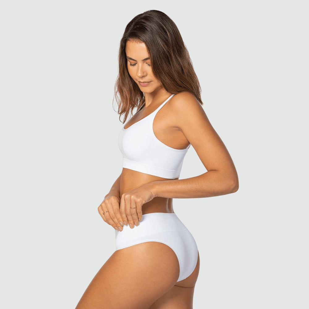 Modelo usando conjunto branco da Cygnuss + Lupo, com top de alças finas e calcinha de cintura alta. Ela está de lado, puxando levemente a peça inferior, demonstrando elasticidade e conforto do tecido.