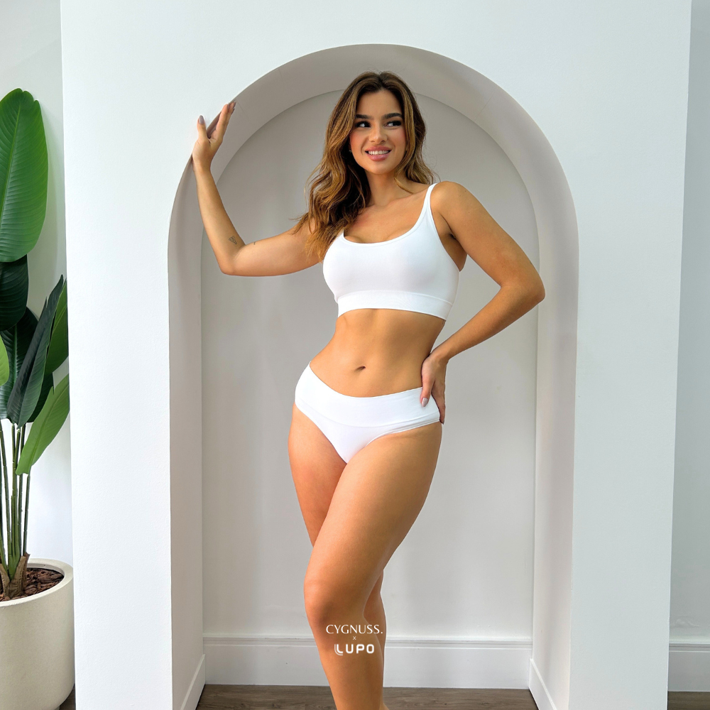 Modelo usando conjunto branco modelador da Cygnuss + Lupo, composto por top e calcinha de cintura alta, posando em pé dentro de um arco decorativo branco com uma mão na parede e outra na cintura. Ambiente iluminado e minimalista.