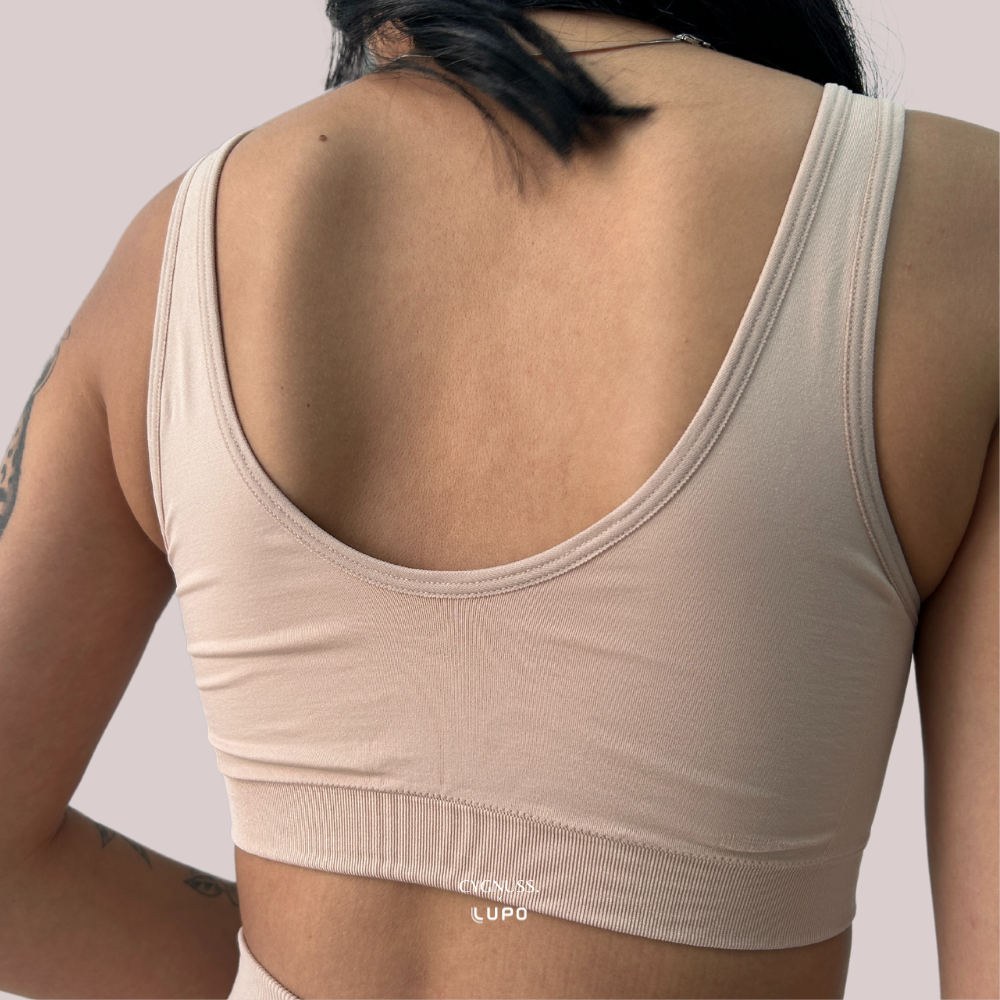 Imagem mostra a parte de trás do top nude sem costura da Cygnuss em parceria com Lupo. A peça possui alças largas e decote profundo nas costas, com cós reforçado para sustentação suave. O fundo em tom rosado claro realça a peça e a silhueta feminina.