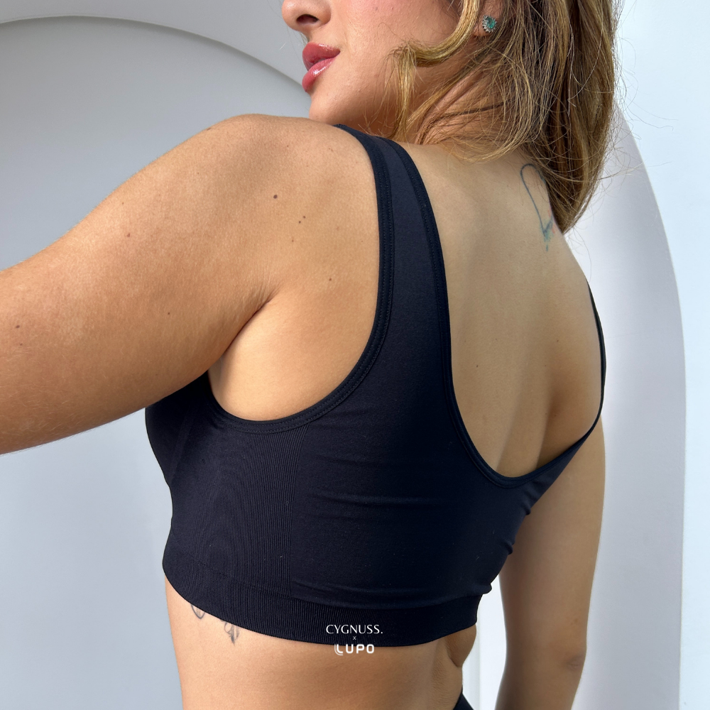 Imagem mostra a parte de trás de uma mulher usando top preto sem costura da marca Cygnuss em parceria com Lupo. A peça possui costas cavadas e cós largo para sustentação. O cenário tem um arco branco moderno que compõe o visual clean e sofisticado da foto.