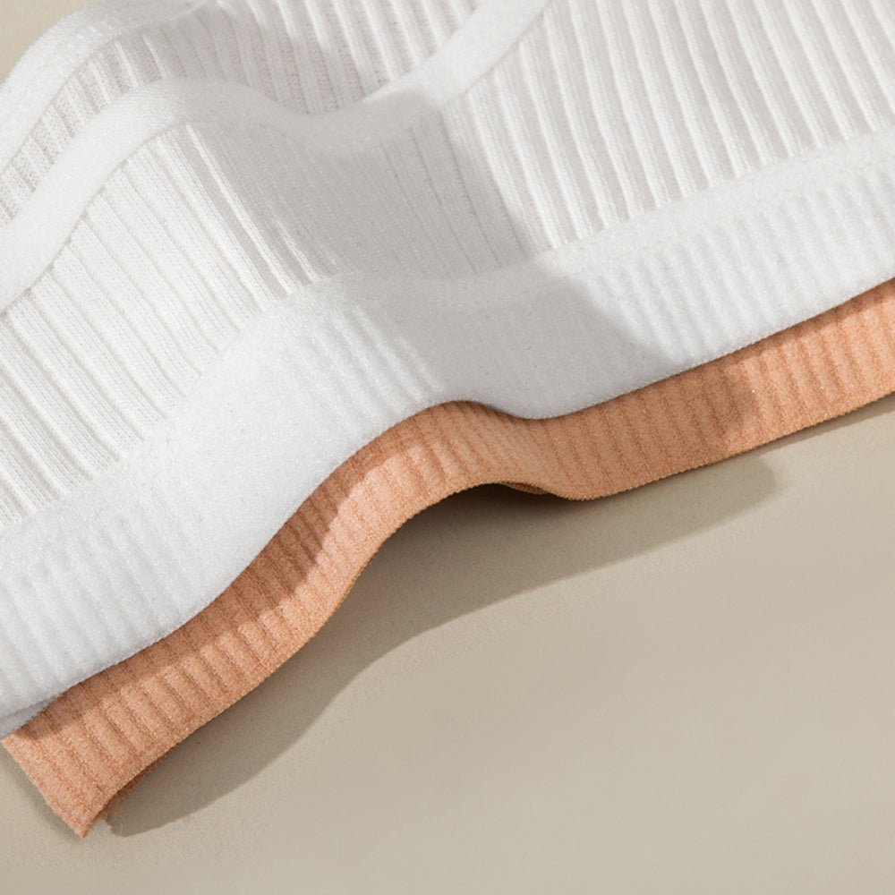 Close em duas calcinhas caneladas sobre superfície clara, nas cores branca e terracota. As peças estão sobrepostas com leve ondulação nas barras, evidenciando a textura do tecido e o caimento leve e flexível do material.