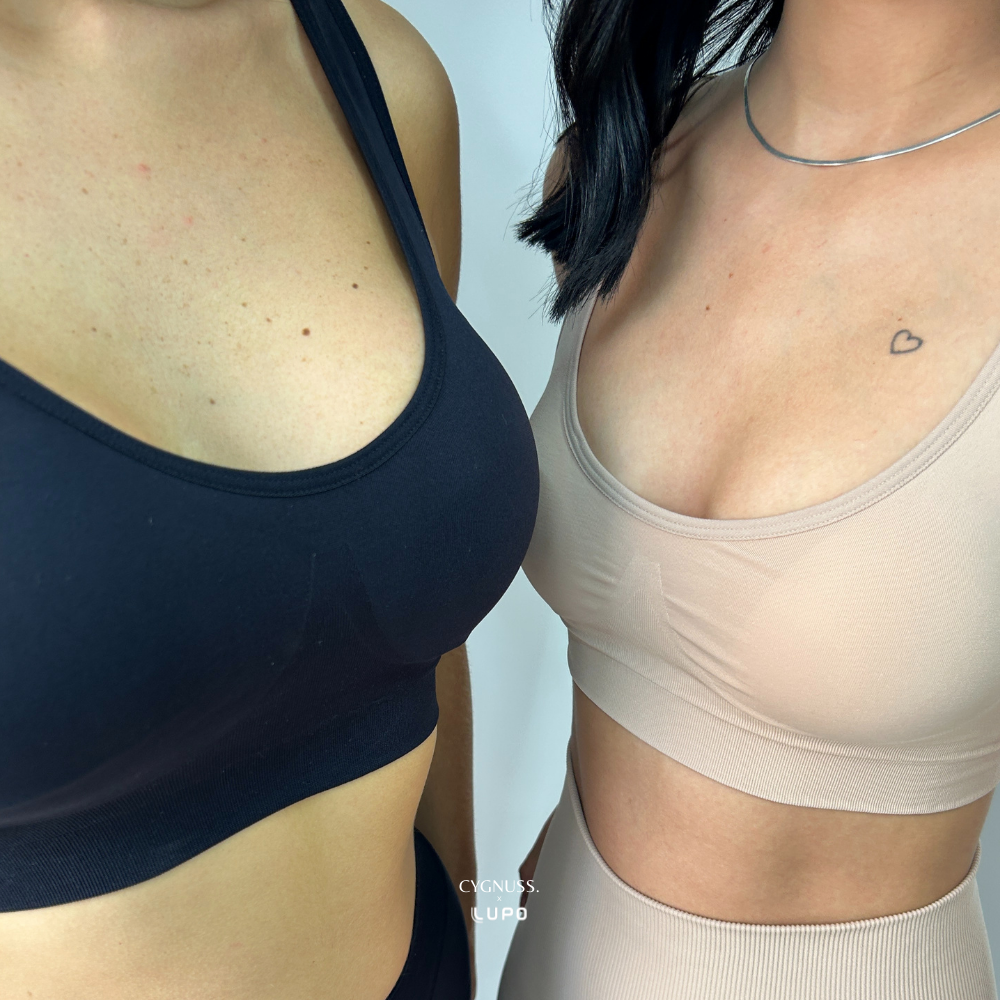 Close frontal de duas mulheres vestindo tops sem costura da Cygnuss em parceria com Lupo. Uma usa o top preto e a outra, o nude. As peças apresentam bojo anatômico, alças largas e modelagem que valoriza o busto com conforto. O fundo claro reforça o contraste entre as cores.