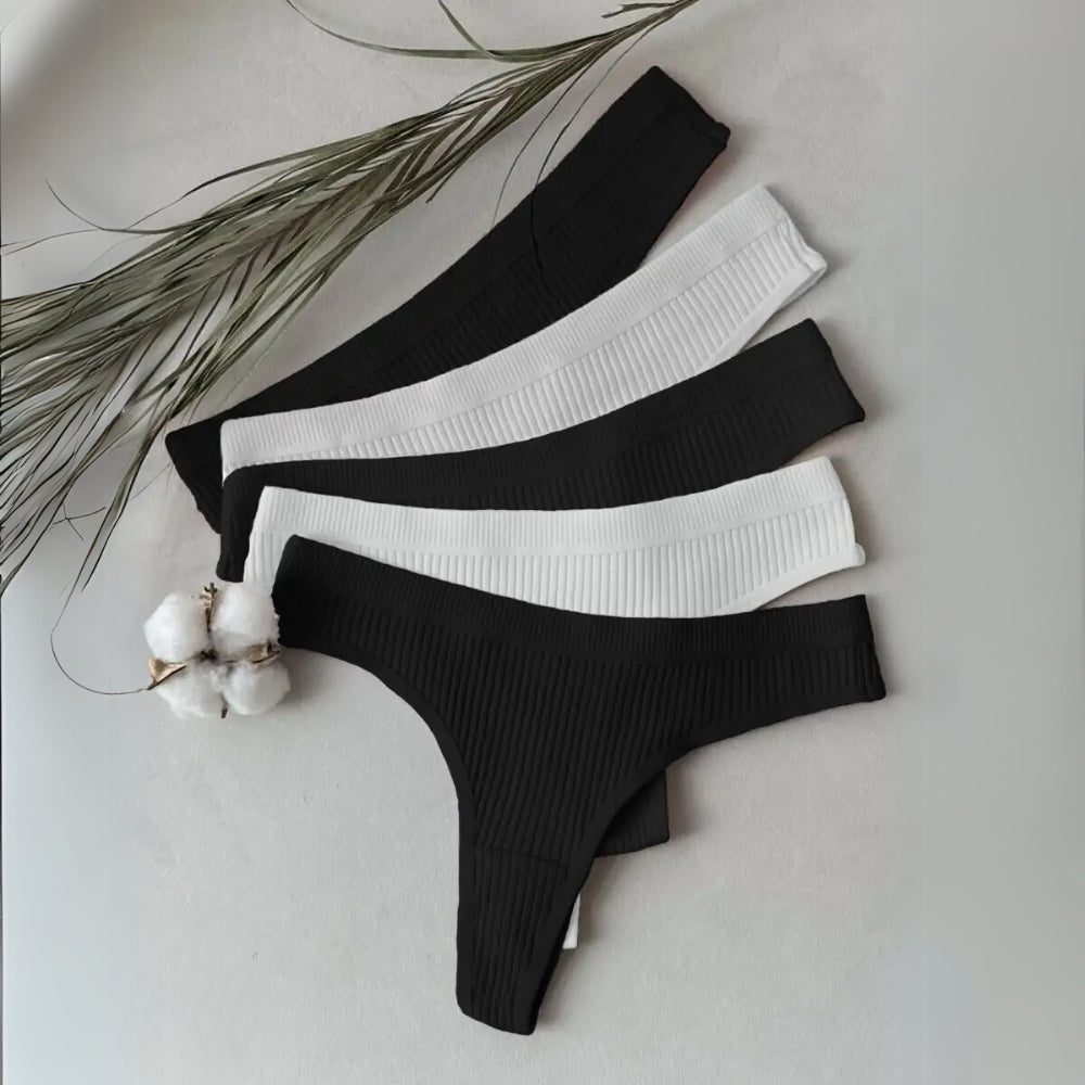 Kit com cinco calcinhas fio dental em tecido canelado, nas cores preta e branca, organizadas em formato leque sobre superfície clara. Ao lado, há ramos secos e flores de algodão, reforçando a suavidade e naturalidade do tecido.