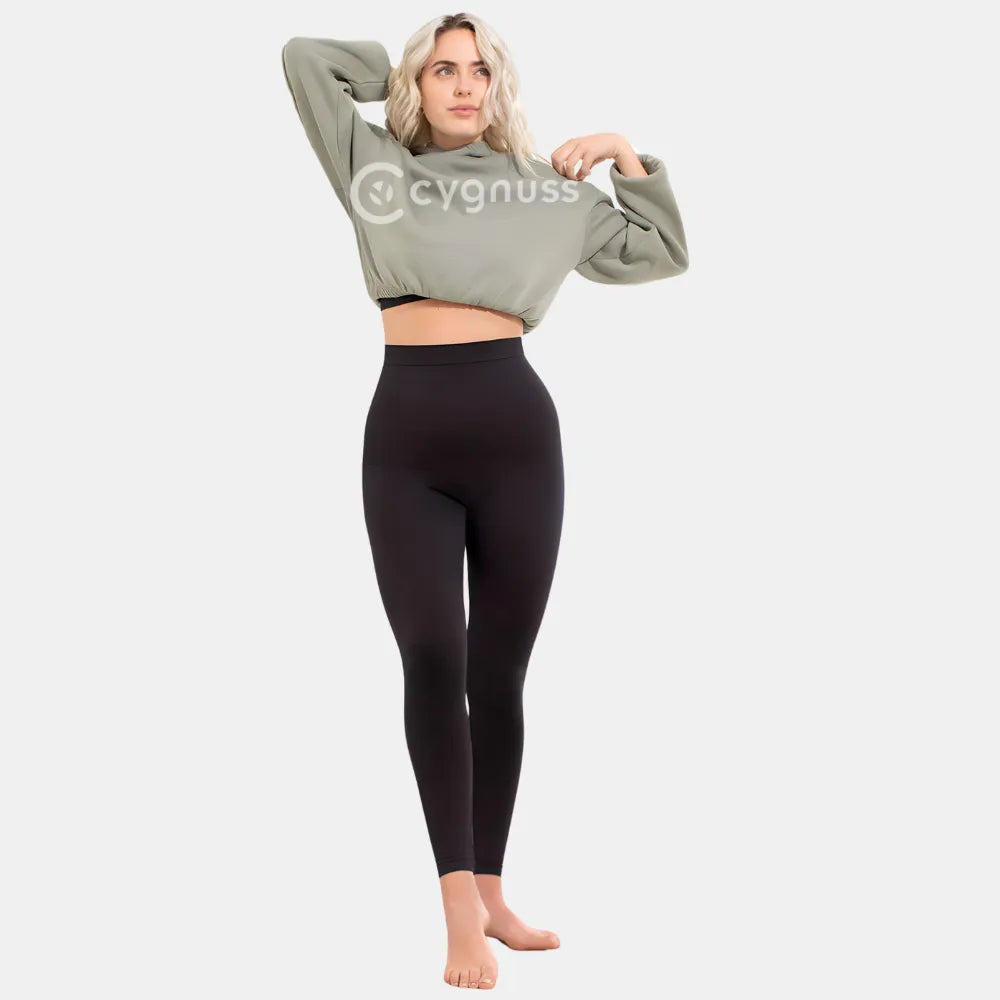 Mulher sorrindo, usando a Legging Jade Fit Modeladora de Cós Alto na cor preta, combinada com hoodie verde, em pose descontraída que destaca o cós alto e o caimento confortável da peça.