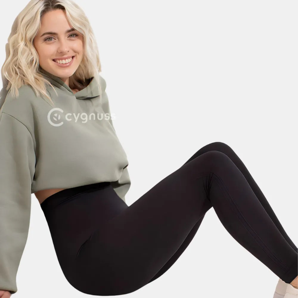 Mulher sorrindo usando a Legging Jade Fit Modeladora de Cós Alto na cor preta, combinada com um hoodie verde, sentada de forma descontraída, destacando o cós alto e o ajuste confortável da peça.